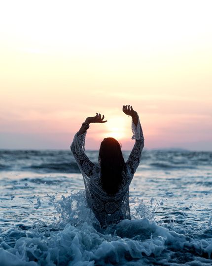 Person standing in ocean waves at sunset with arms raised toward the sky