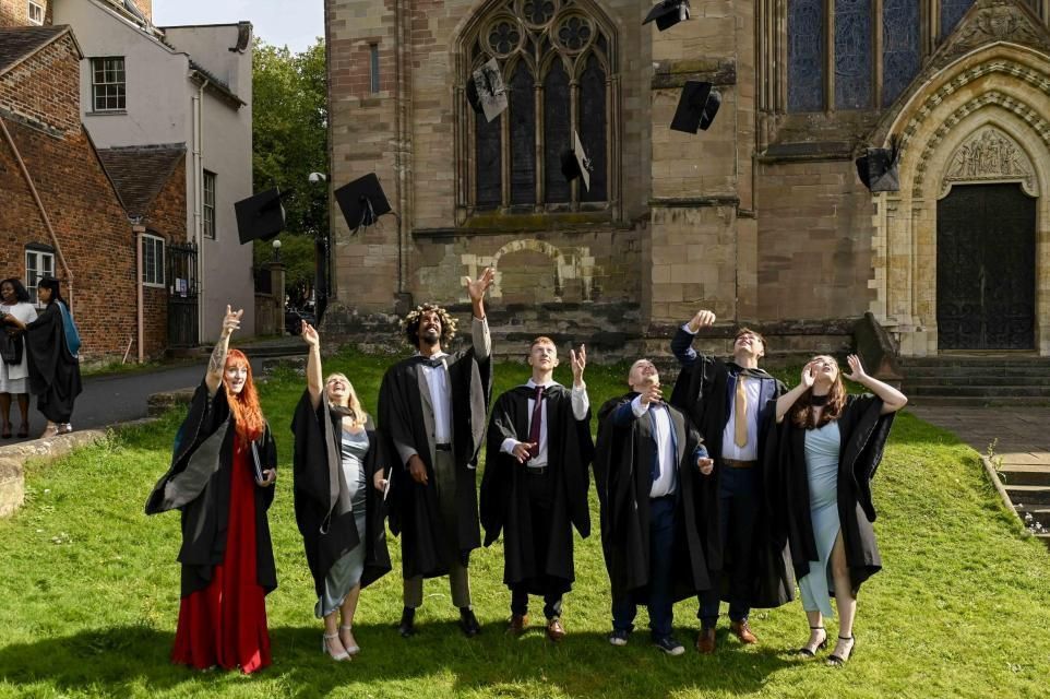 Seven graduates in caps and gowns pose on a lawn outside a stone church.