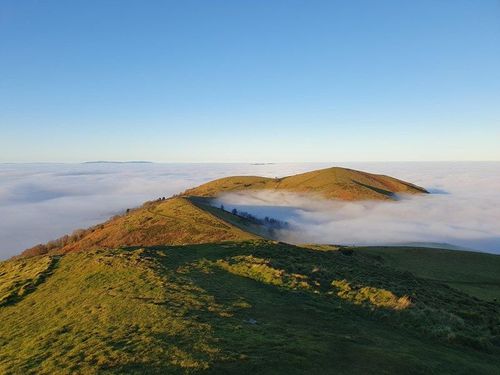 A grassy ridgeline in Malvern Hills rising above a thick layer of clouds under a clear blue sky.