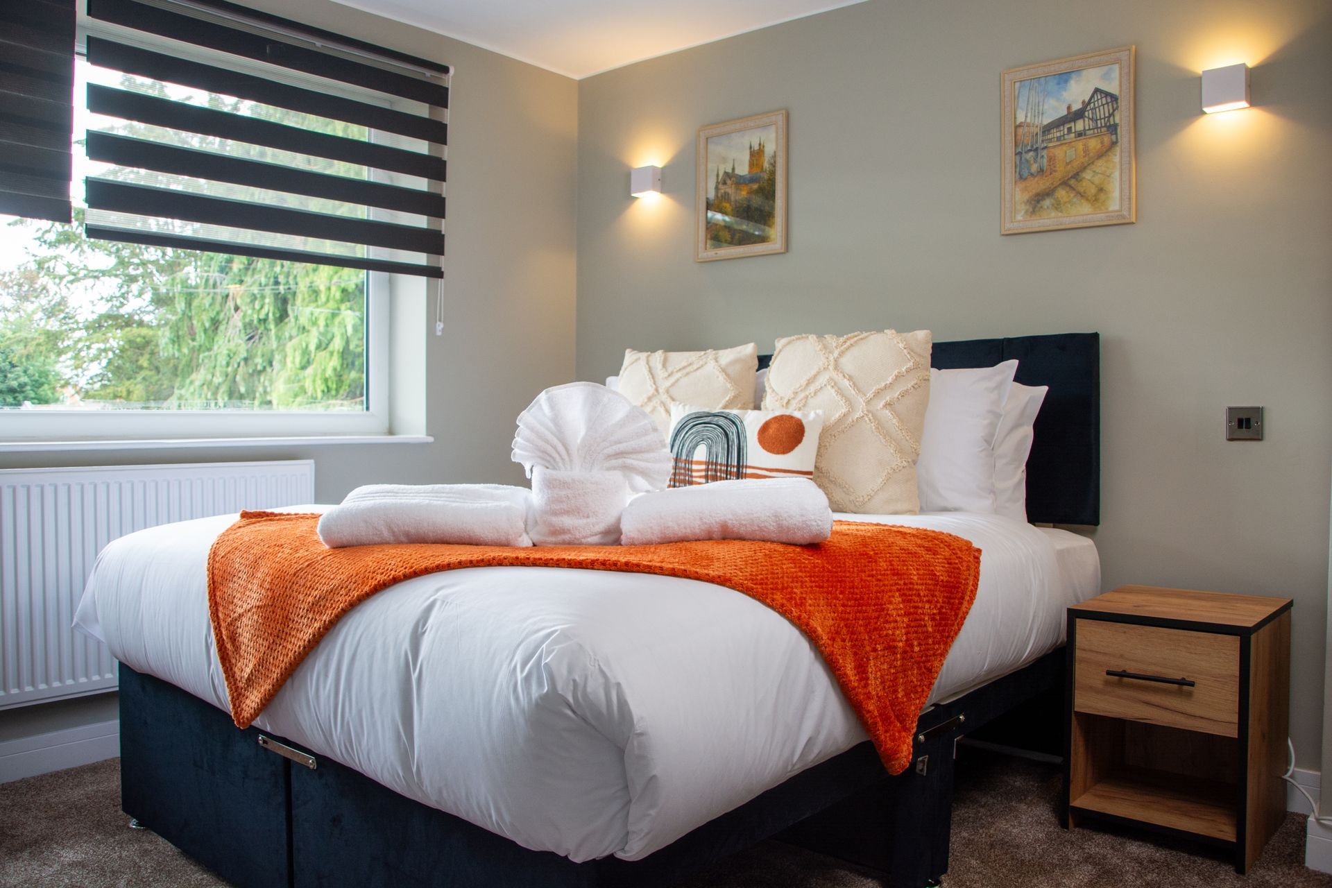 Modern bedroom with a neatly made bed, orange throw, striped blind, and two framed pictures on gray walls