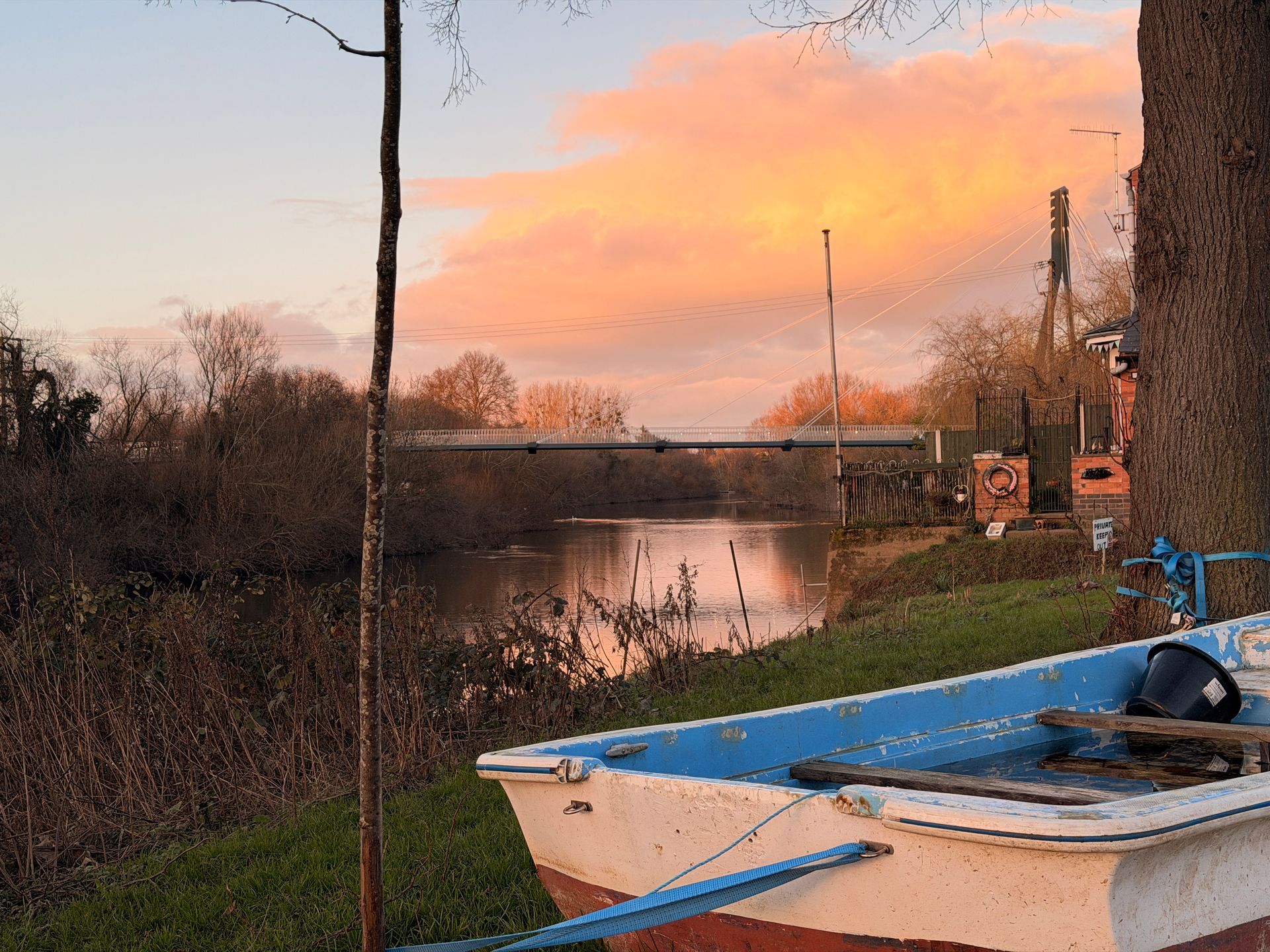 Small boat by a calm river at sunset, with pink sky and bare trees along the bank.