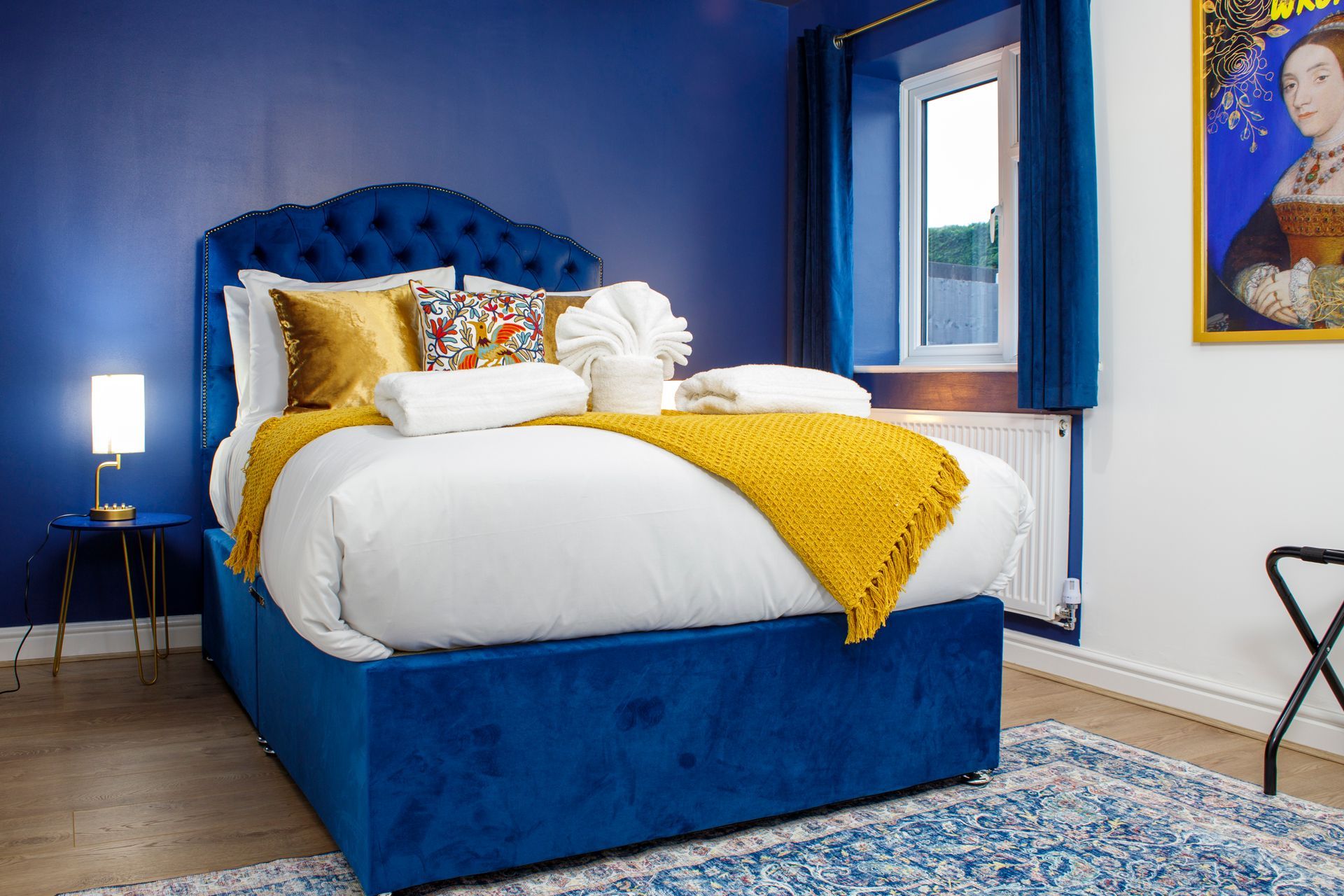 Blue bedroom with a tufted bed, white and yellow bedding, blue curtains, and a patterned rug.