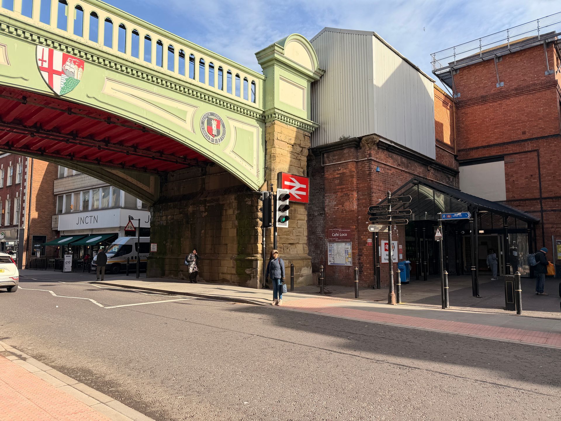 Street view under a green railway bridge beside brick buildings and a pedestrian walkway.