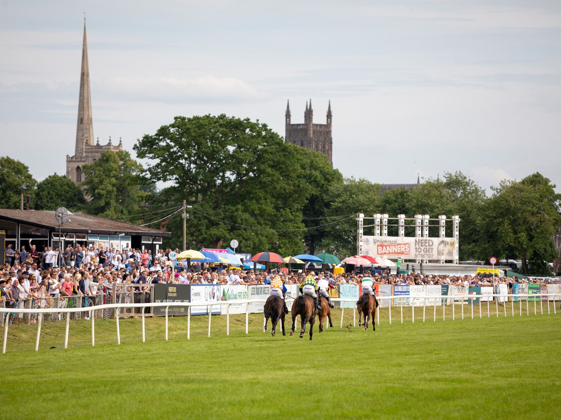 Horse race on a green field with spectators, white rails, and a cathedral skyline in the background