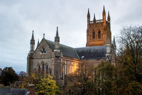 A historic Worcester cathedral with a tall, ornate stone tower, shown against a cloudy sky surrounded by autumn trees.