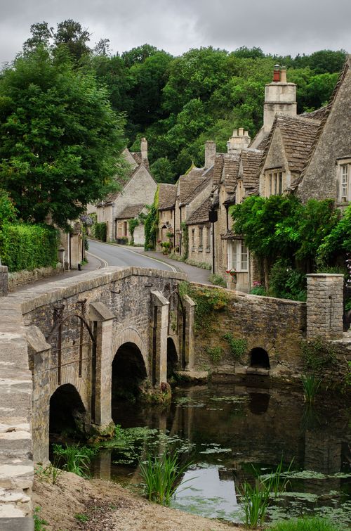 Stone bridge over a narrow stream beside thatched cottages in a green village.