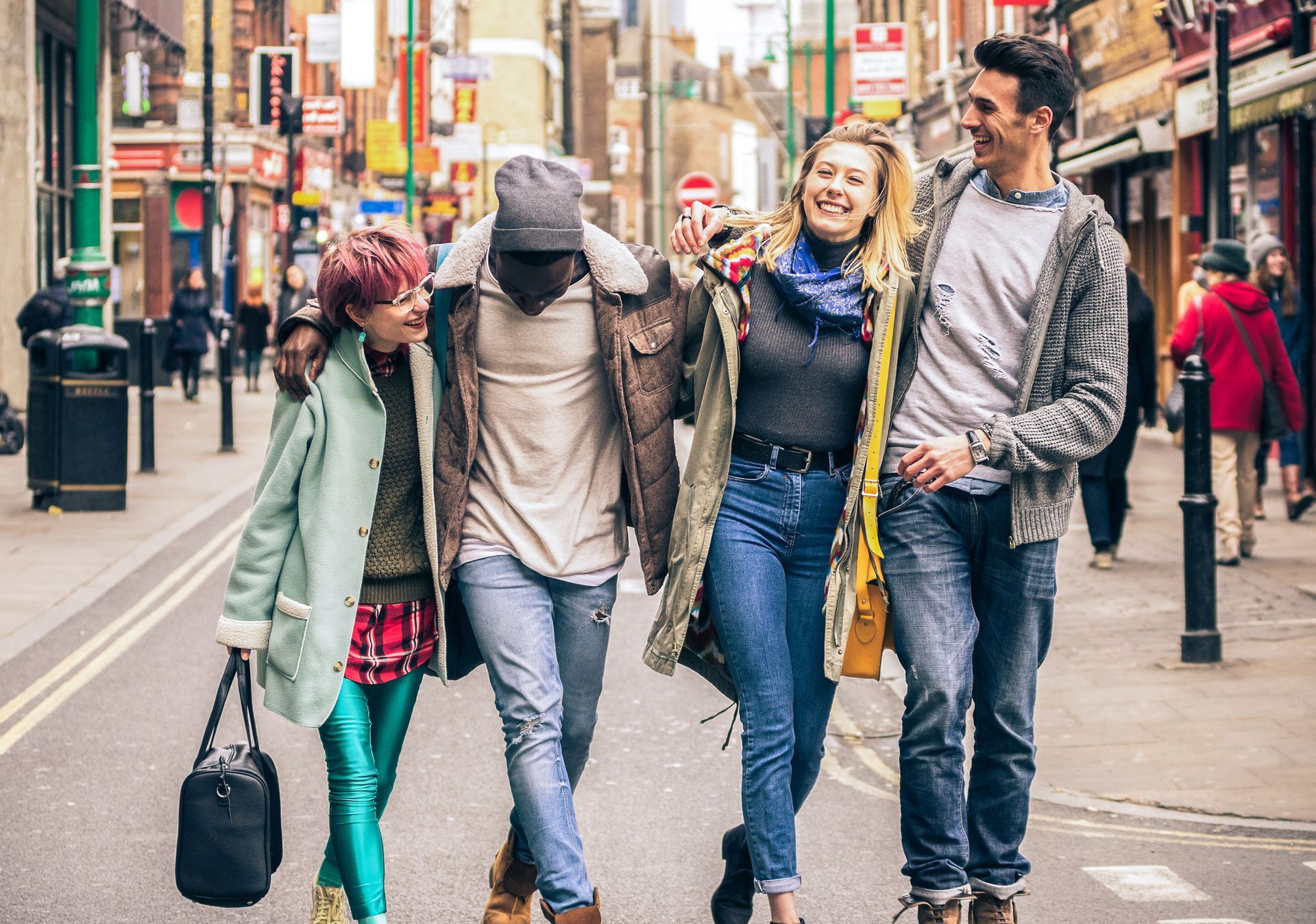 Four friends walking arm-in-arm down a city street, smiling and chatting.