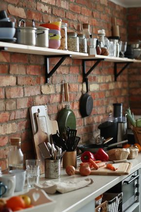 A kitchen with rustic brick walls, open shelves holding jars and cookware, and a counter with fresh vegetables and tools.