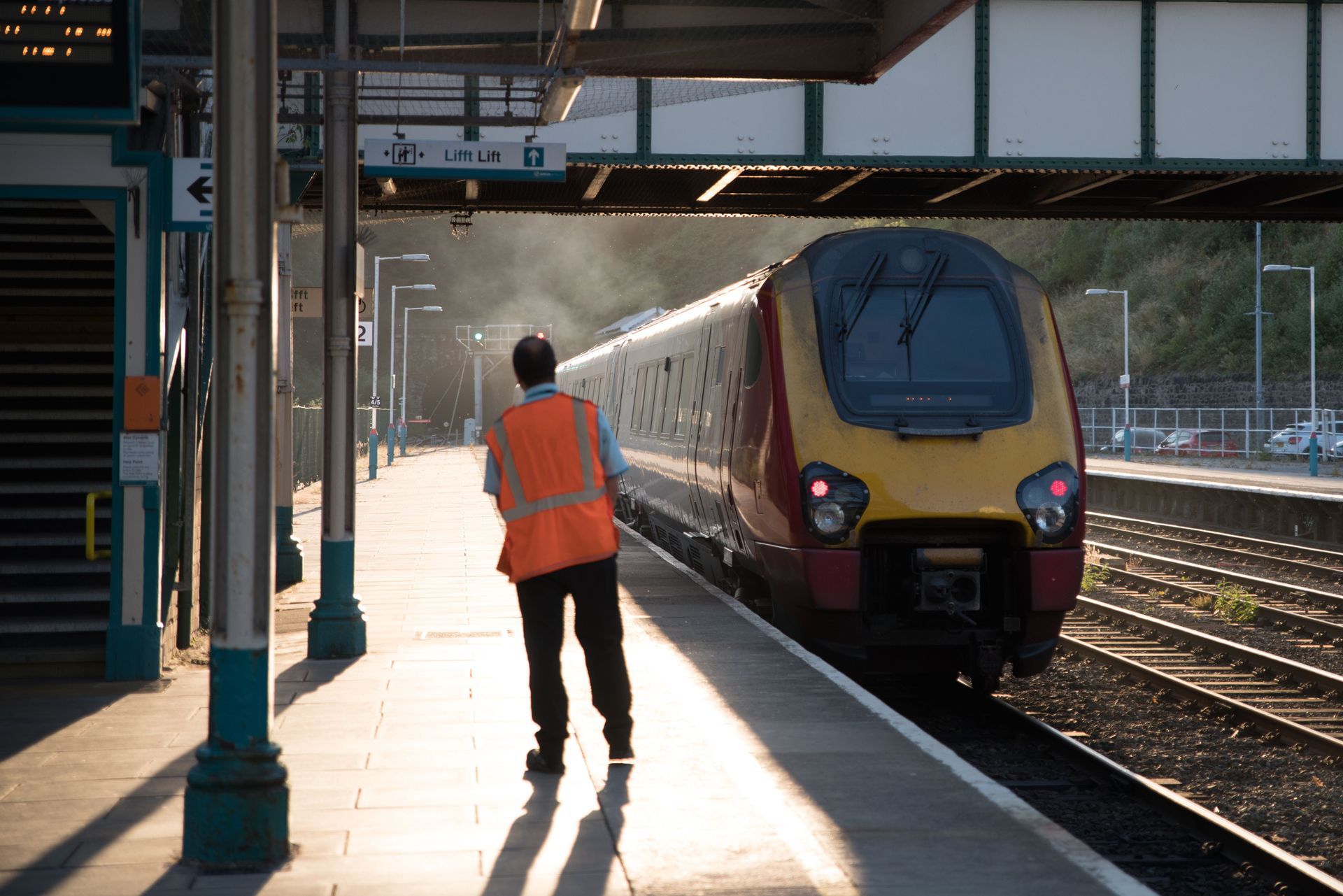 Railway platform with a worker in orange vest signaling a yellow train arriving at sunset
