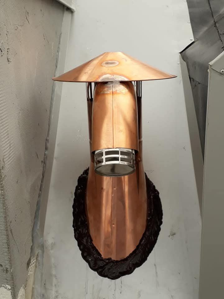 Griglia di ventilazione a muro in rame con cappuccio conico e finiture scure, montata su una parete esterna grigia.