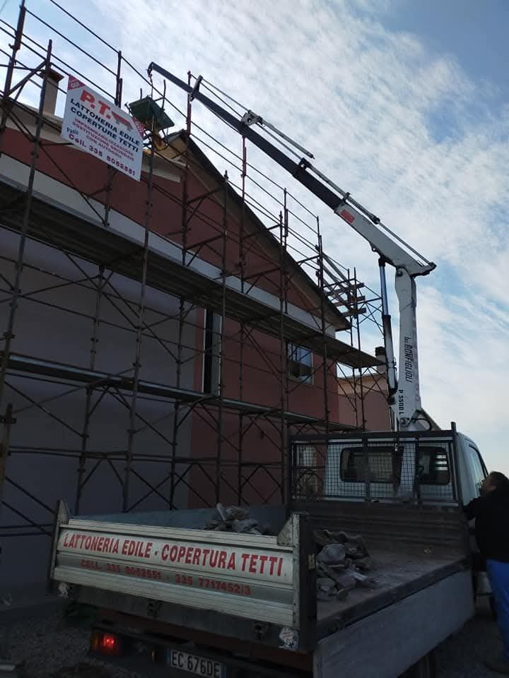 Un operaio su un'impalcatura accanto a un camion che solleva materiali verso un edificio con il tetto rosso.