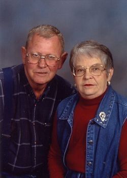 Two adults posing together against a blue studio backdrop, wearing glasses and casual clothing.