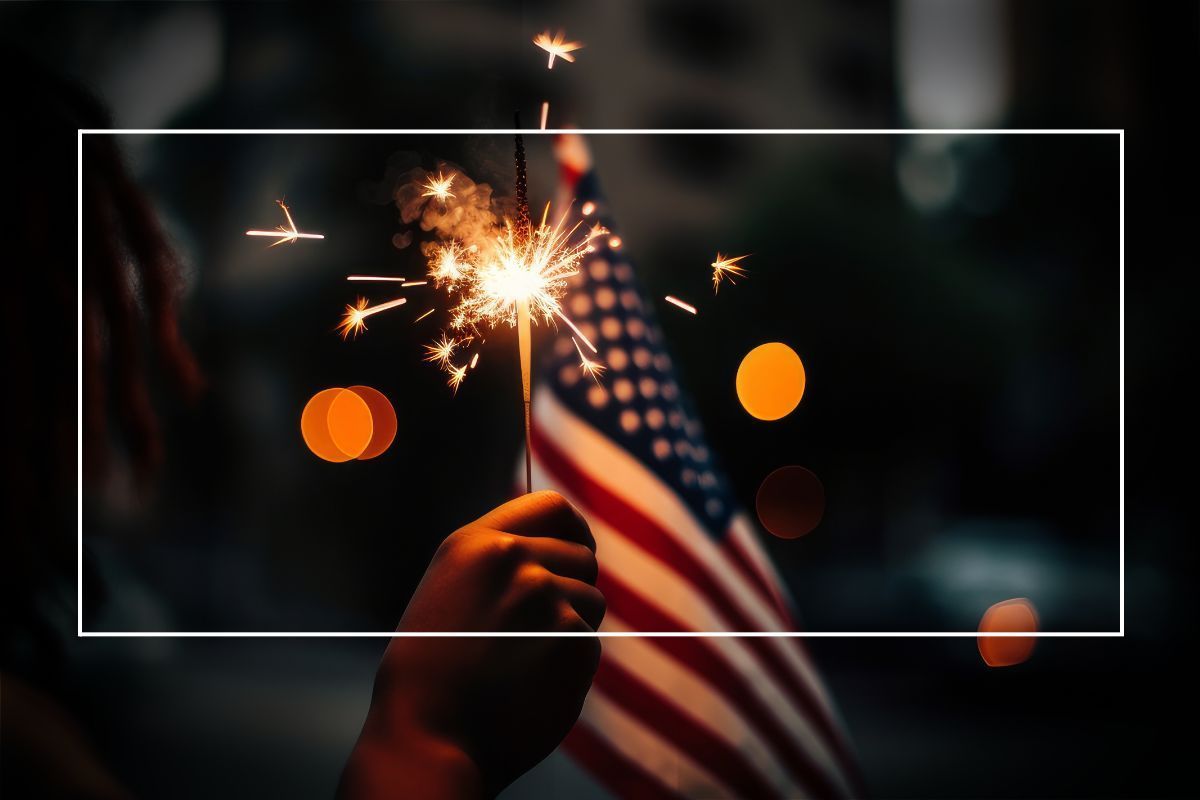 Hand holding a sparkler beside a small American flag at night with blurred lights in the background
