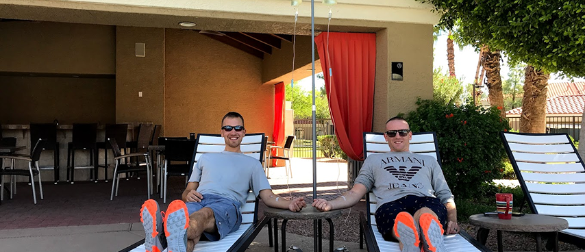Two men receiving IV drip therapy while relaxing on outdoor lounge chairs