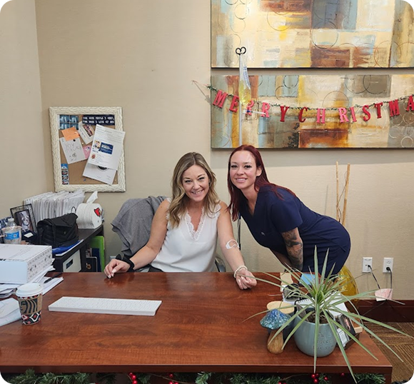 Two women sitting at a desk with a merry christmas banner hanging on the wall behind them