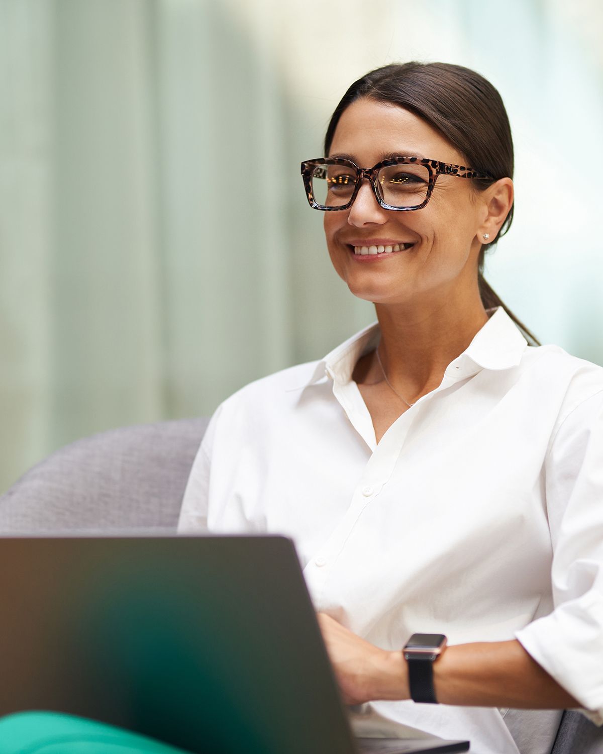 Smiling professional in glasses and white shirt, seated beside a laptop in a bright office setting