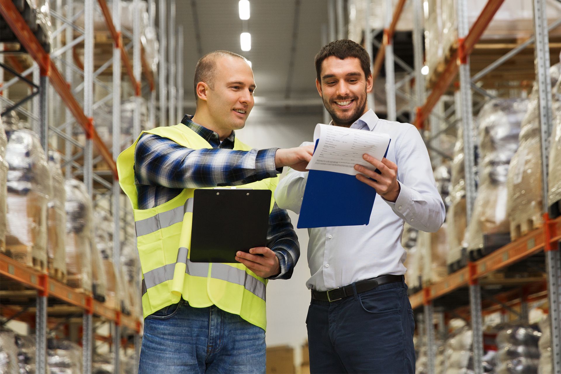 Two warehouse workers in safety vests review papers amid tall storage racks.