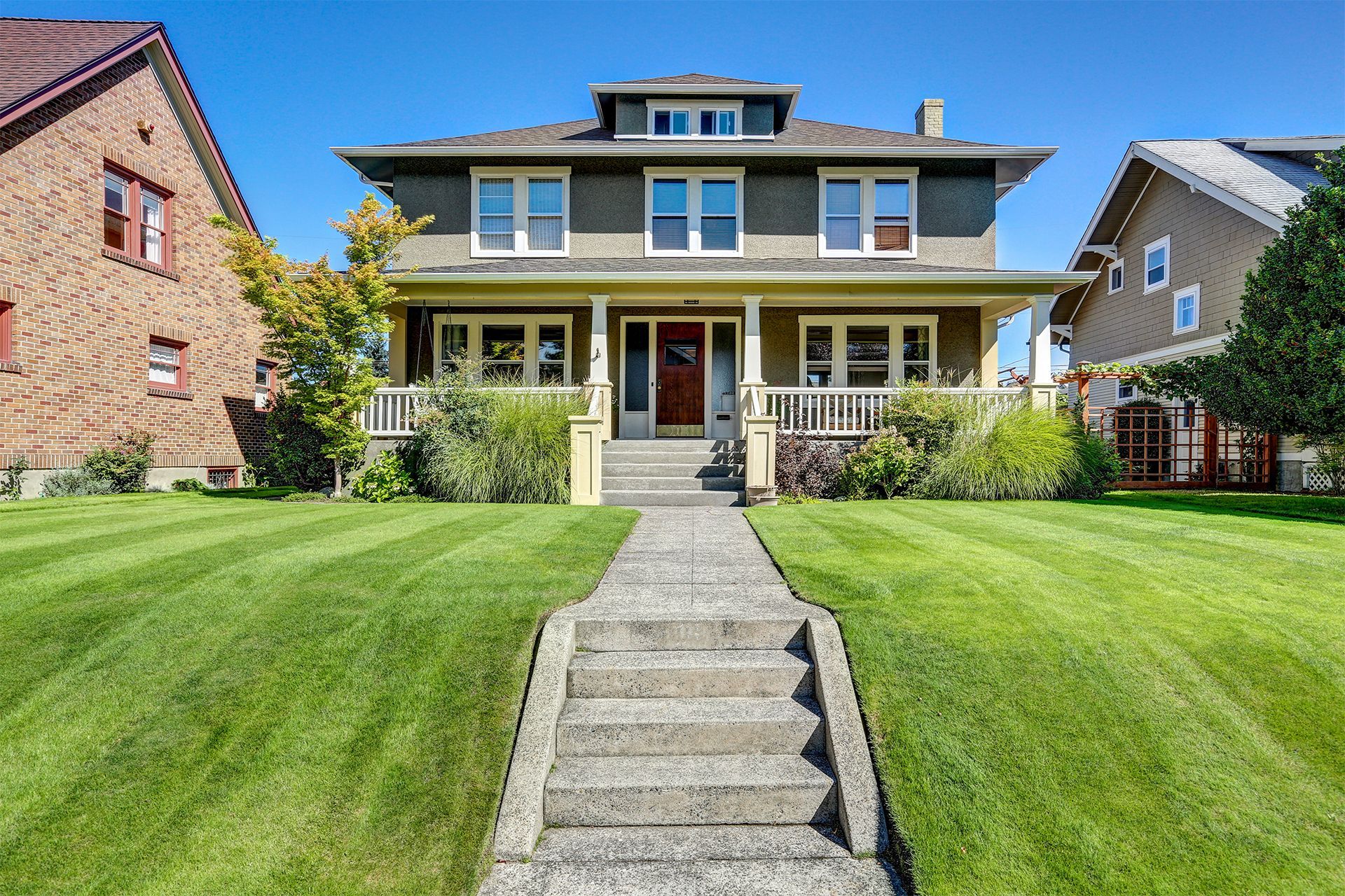 Large two-story house with a front lawn, stone steps, and a central porch under a clear blue sky.