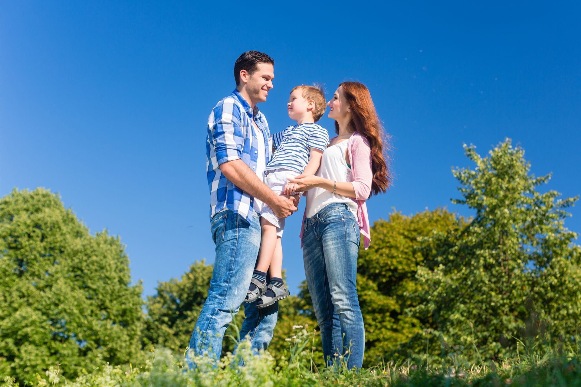 Three people standing in a grassy park under a bright blue sky, looking at each other and smiling.