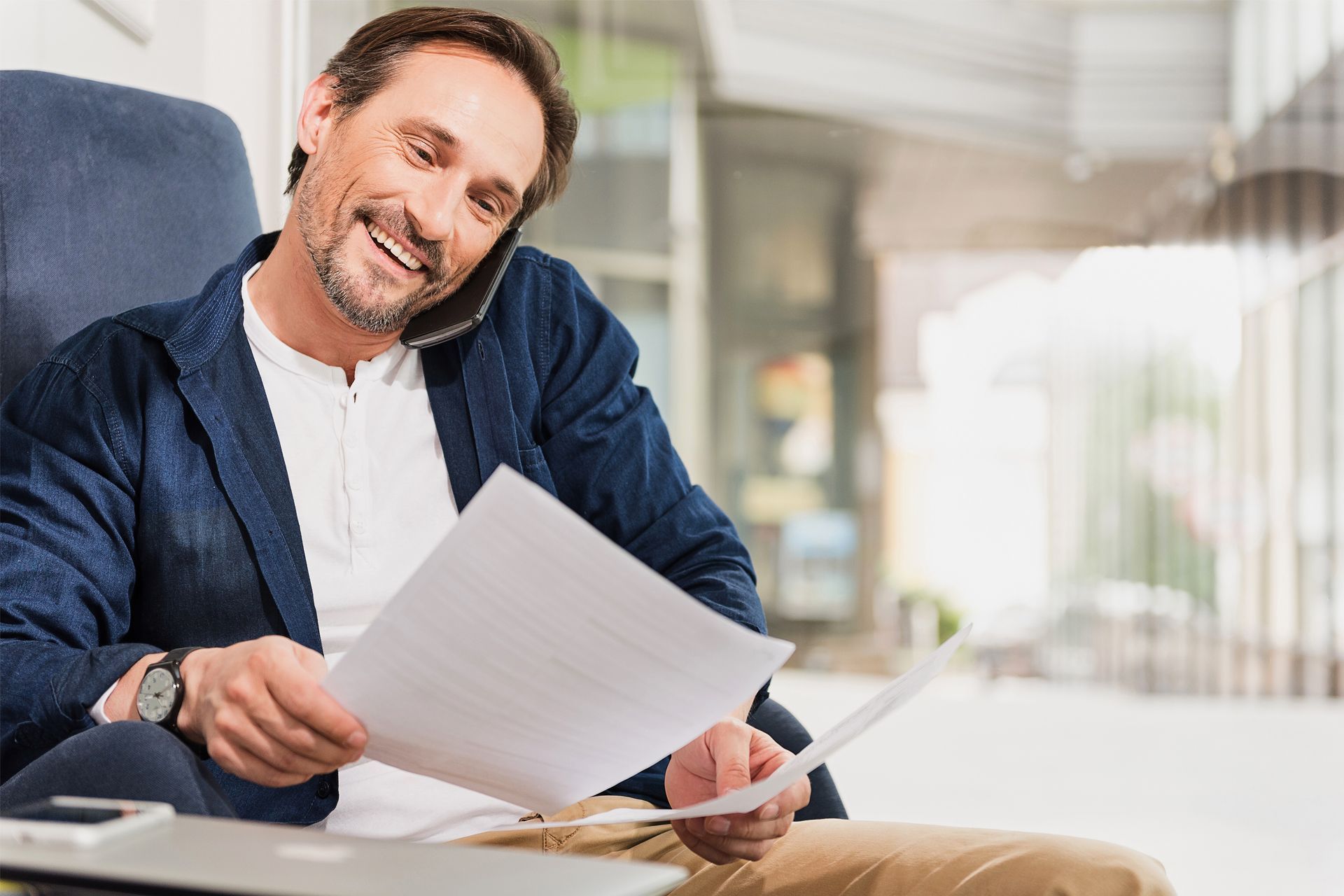 Man smiling on a couch, reading paperwork with a pen in hand in a bright living room.