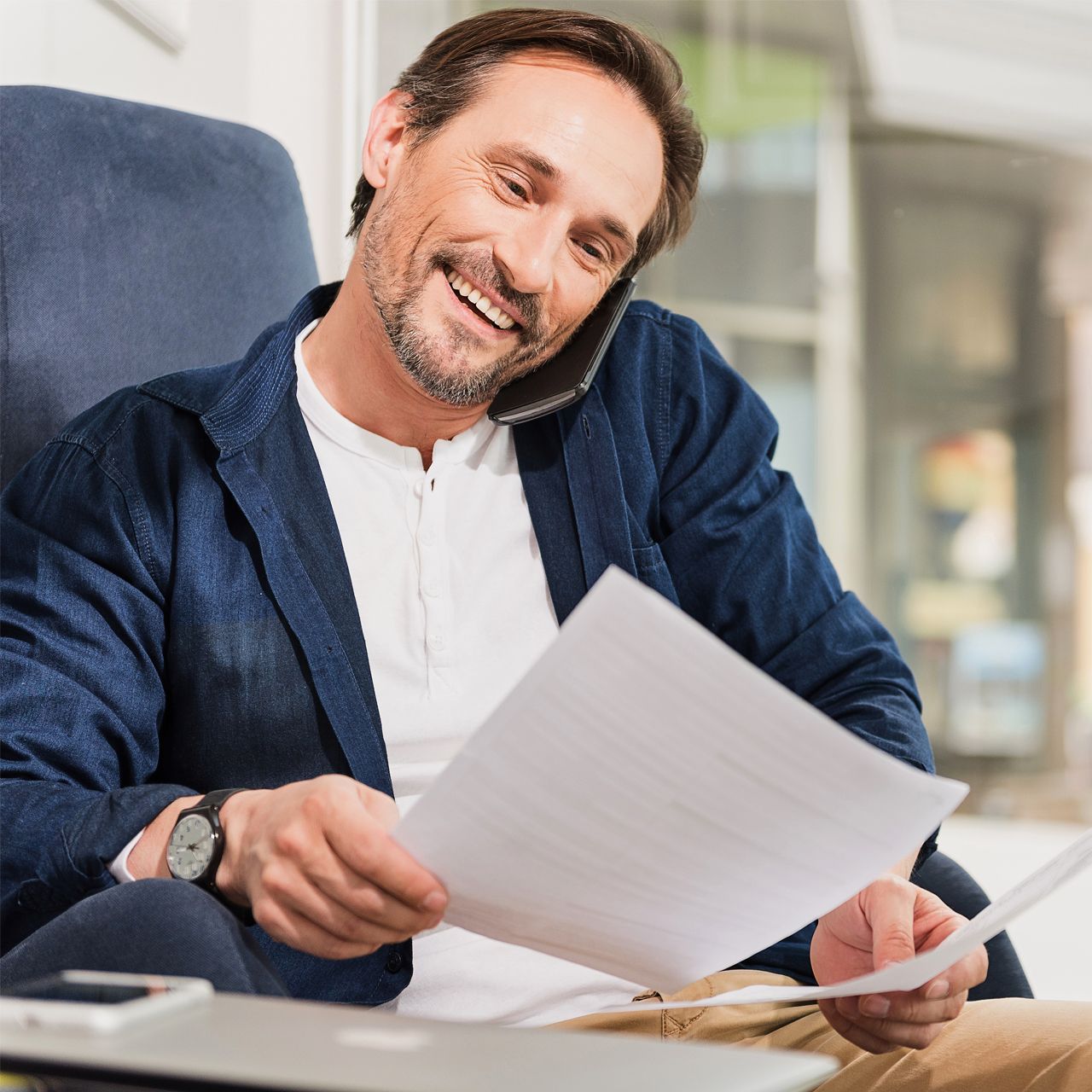Smiling man in a navy blazer reviewing papers at a desk in a bright office.