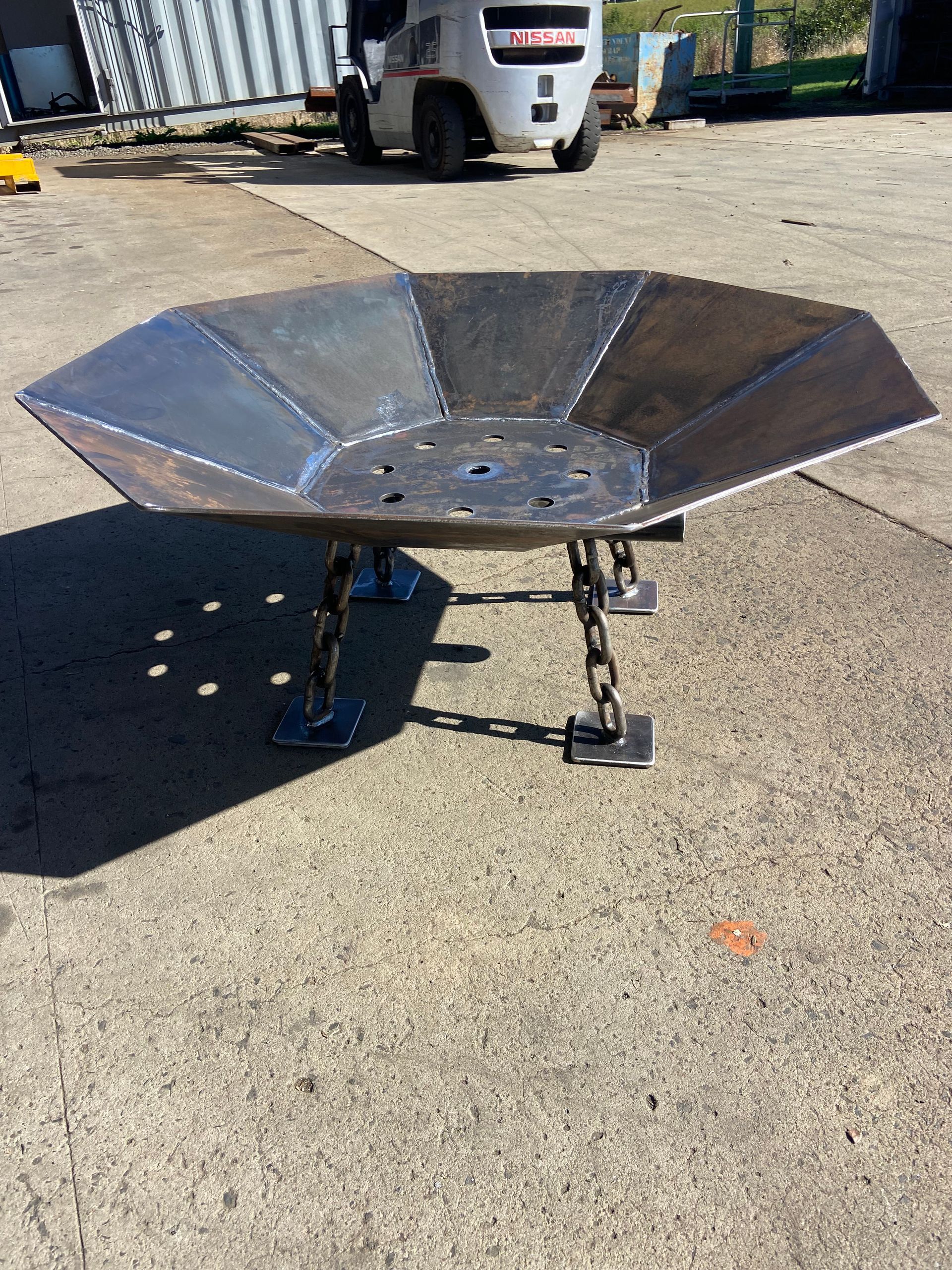 Steel fire pit with octagonal bowl, suspended by chains and metal legs, outdoors on concrete.