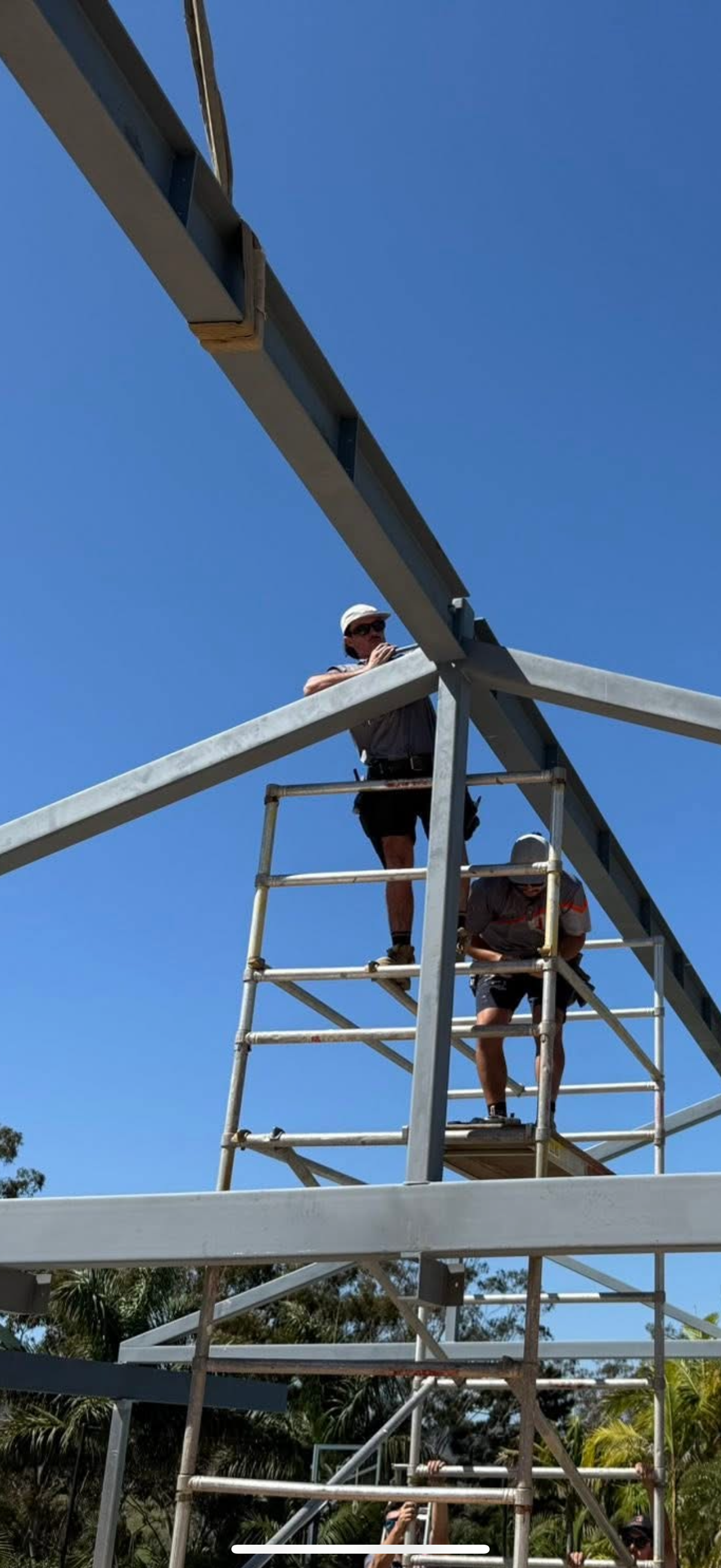 Two construction workers on scaffolding, assembling a steel frame outdoors against a blue sky.