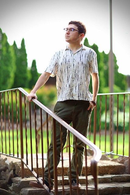 Man in glasses stands on steps, hand on railing, looking upward. Wearing a patterned shirt and olive pants.
