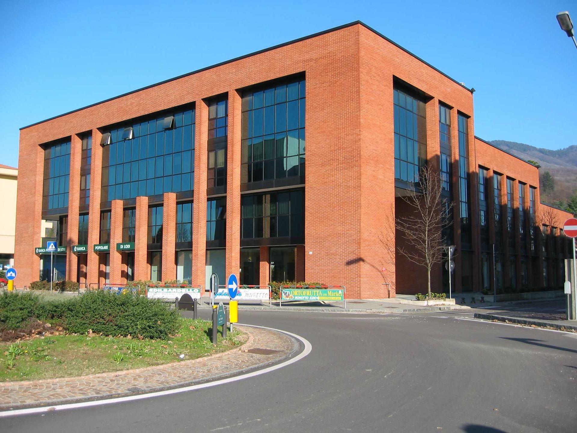Moderno edificio per uffici in vetro con finestre blu riflettenti e colonne rosa-beige, incorniciato da alberi spogli.