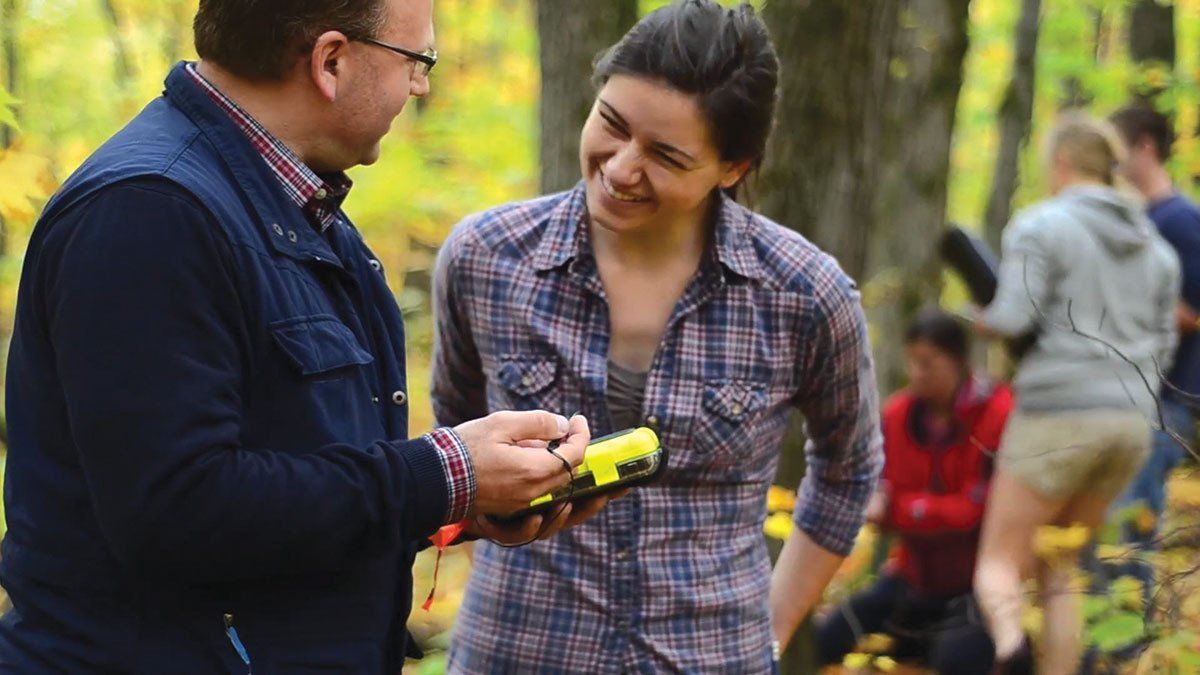 A man and a woman are standing in the woods talking to each other.
