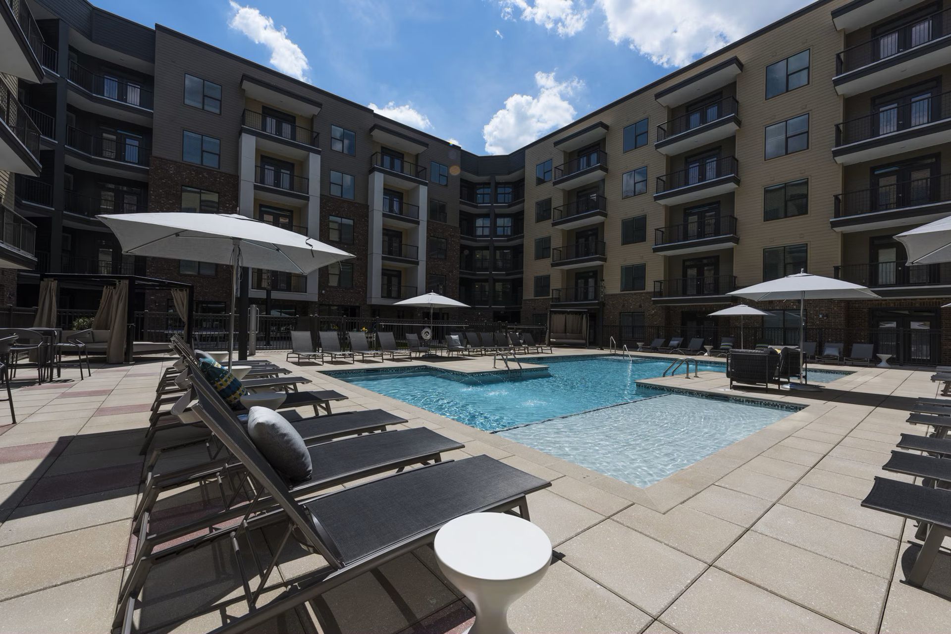 Apartment complex pool area with lounge chairs, umbrellas, and blue water under a sunny sky.