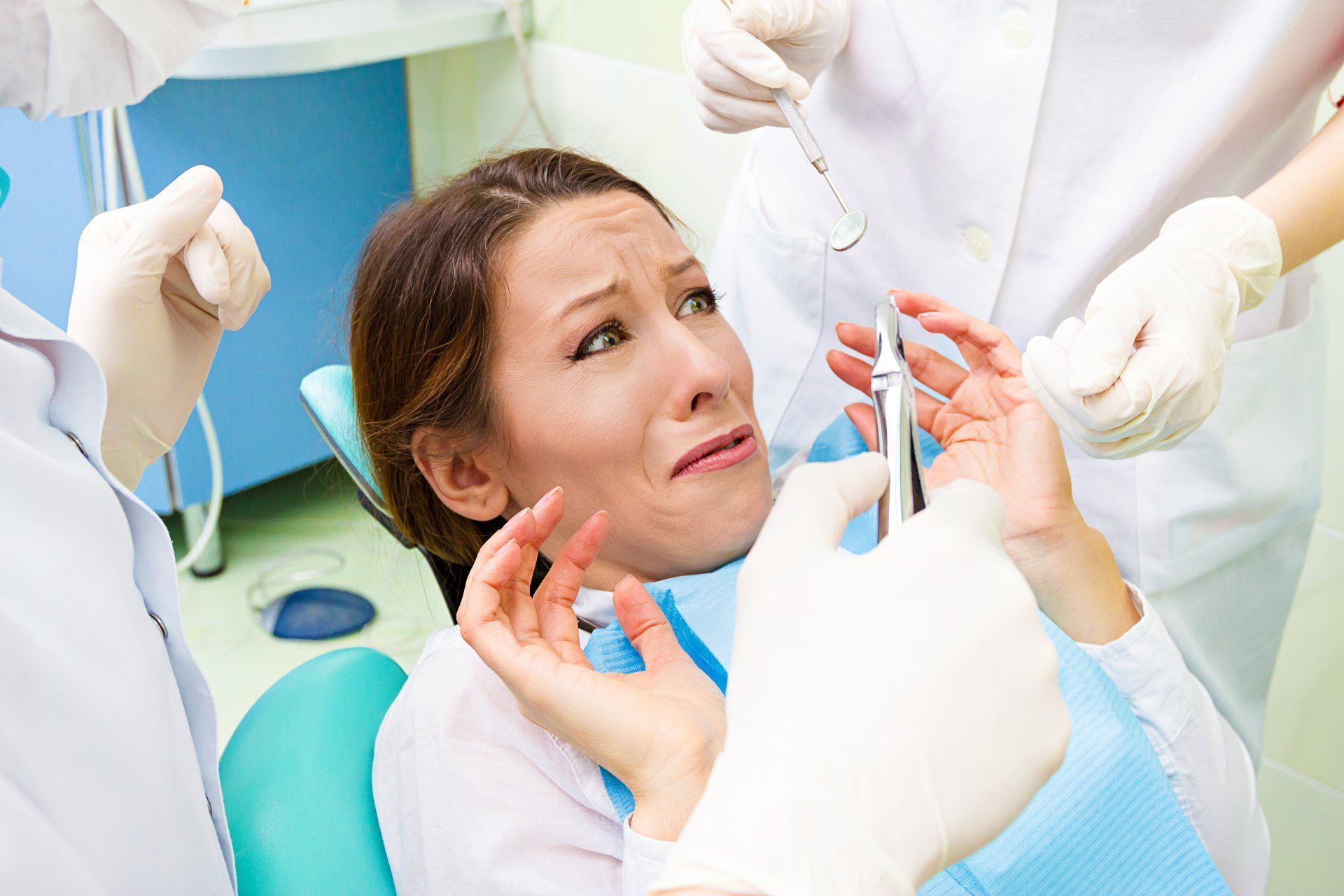 Woman in dental chair, scared, multiple gloved hands holding instruments, bright clinic setting.