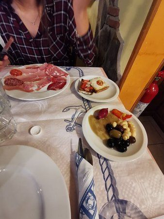 Piatti di affettati e frutta su un tavolo di ristorante, accanto a un cliente seduto che tiene in mano un telefono.
