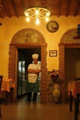 Lo chef è in piedi sulla soglia di un ristorante rustico, illuminato da una luce calda, con archi in mattoni e tavoli apparecchiati.