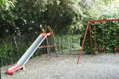 Parco giochi vuoto con scivolo rosso e altalena su ghiaia, circondato da alberi.