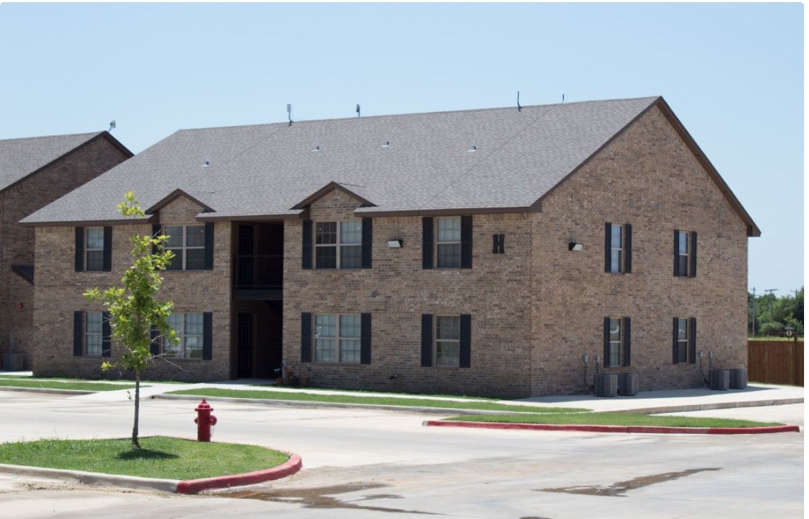 Two-story tan brick apartment building with a small tree and parking lot in front