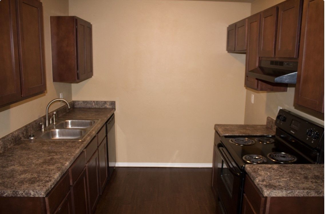 Empty kitchen with dark wood cabinets, brown countertops, double sink, and black stove.