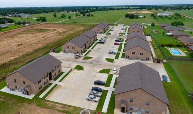 Aerial view of a suburban apartment complex with parking lots, cars, and green fields nearby