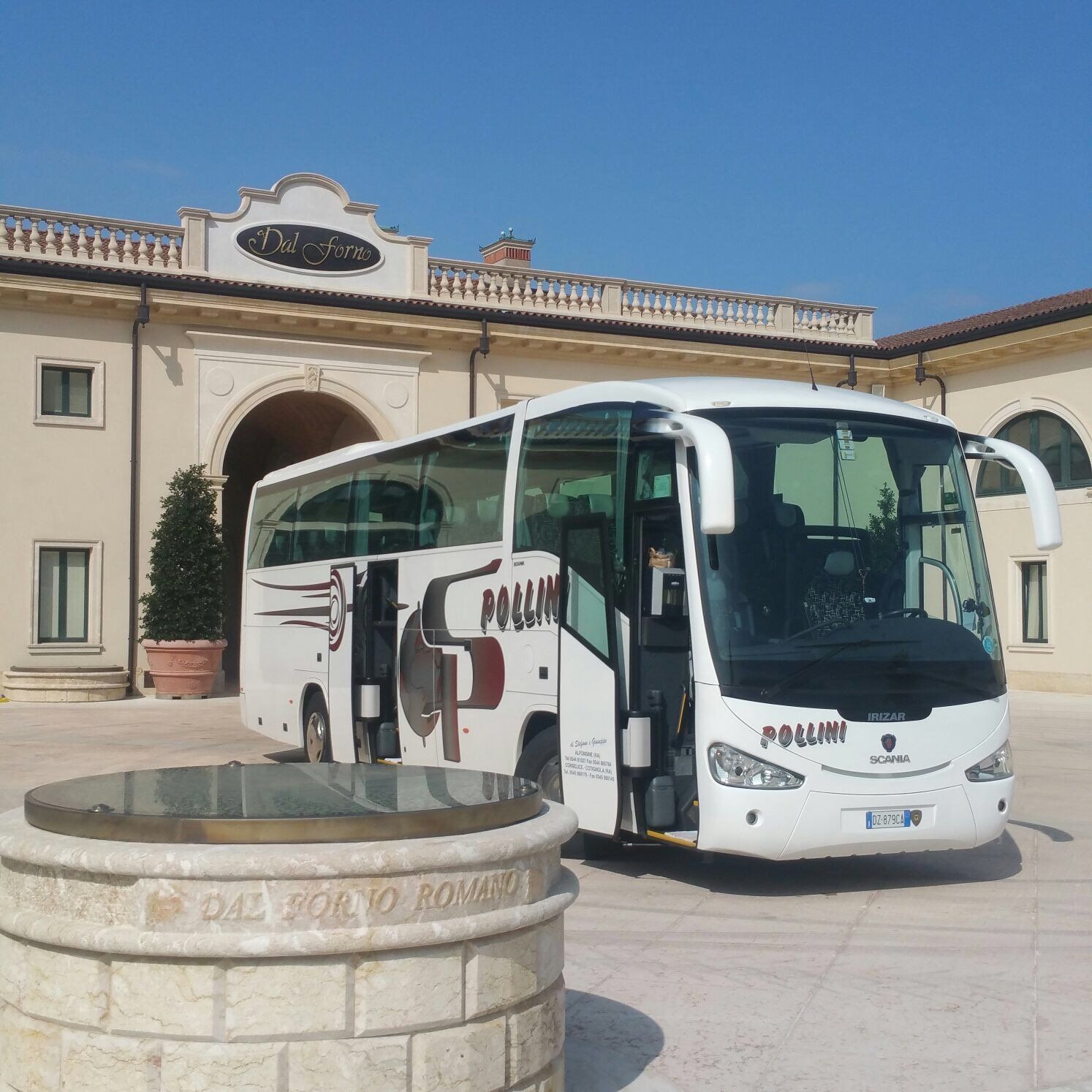 A white tourist bus parked outside a building with arched entrances. In the foreground is a well.