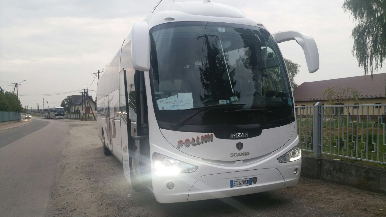 A white Pollini bus parked on a road, with the door open. Buildings and sky in the background.
