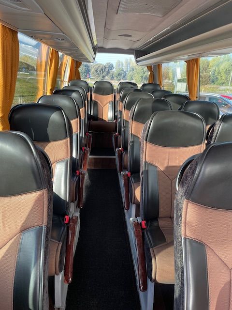 Interior of a tourist bus with rows of black and beige seats, a central aisle and windows with yellow curtains.