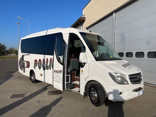 A white Polla bus with its door open, parked outside a building on a sunny day.