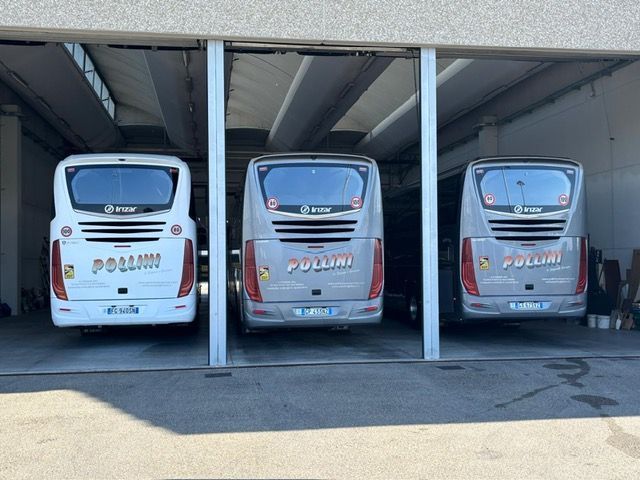 Three Pollini buses parked next to each other under a covered area. The buses are grey and white.
