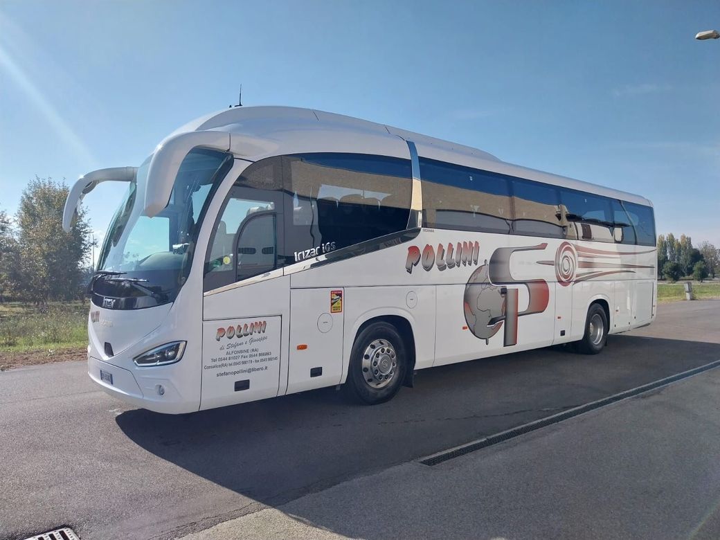 A white bus parked near a building with an archway. In the foreground is a well.