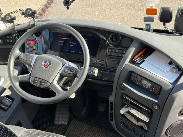 Interior of the driver's cab of a Scania bus, showing the steering wheel, the dashboard with controls and the windscreen.