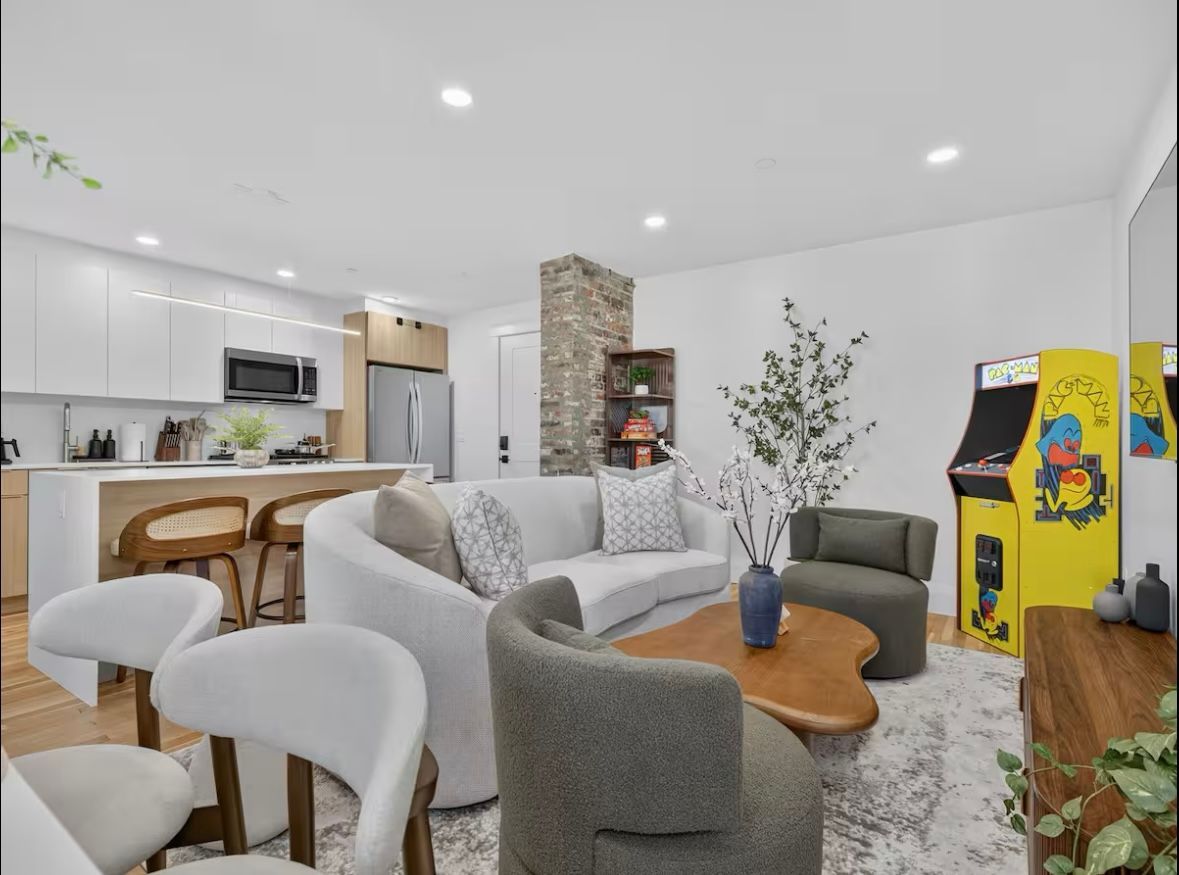 Living room with white walls, kitchen, arcade game, curved sofa, chairs, and wooden coffee table.