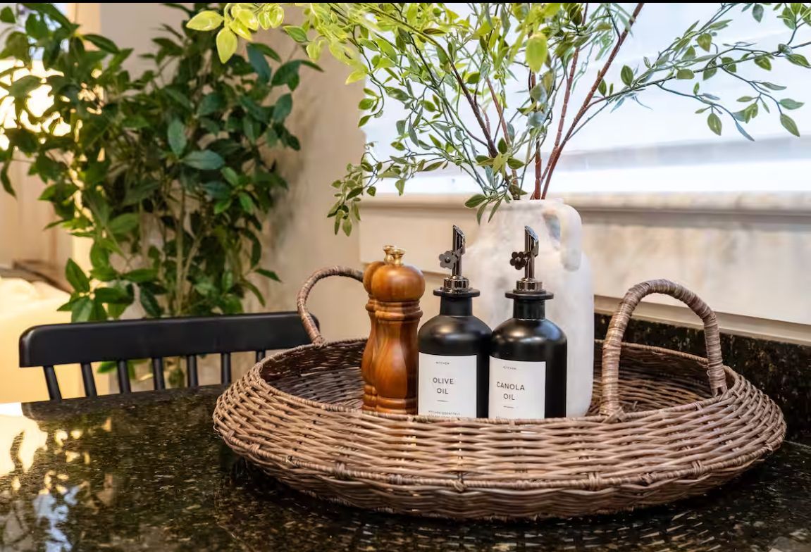 Wicker tray with salt and pepper grinder, oil bottles, and vase with greenery on a countertop with a plant.