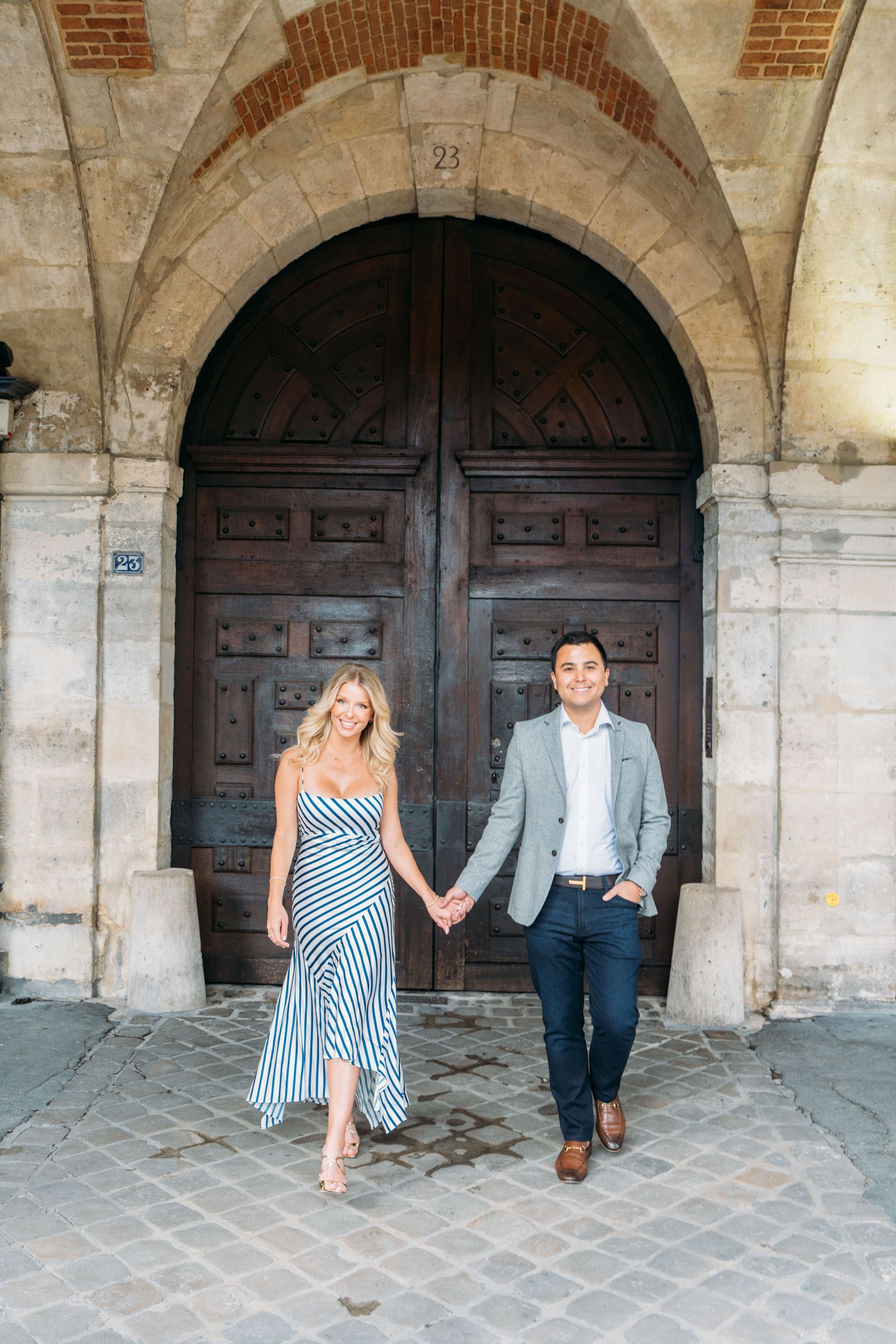 Couple, walking in front of a large arched doorway. She wears a blue and white patterned dress, he wears a blazer.
