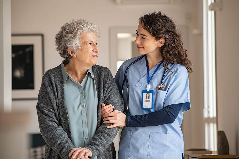 Nurse assists elderly woman with walking indoors; both smiling.