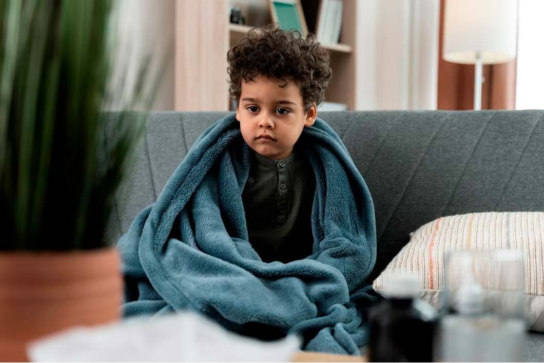 Boy wrapped in a blue blanket, looking unwell. Sitting on a couch, home interior setting.